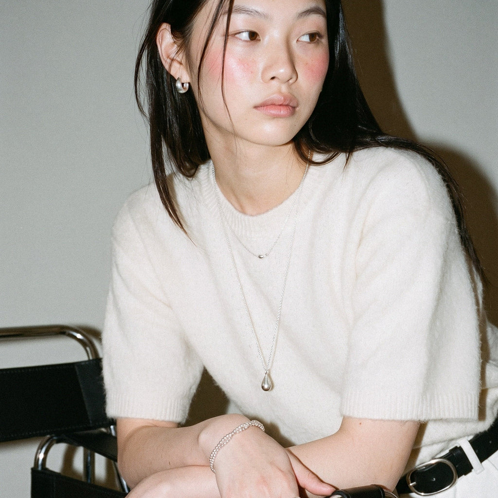 A woman wearing a cream-colored short-sleeve knit sweater and white pants, accessorized with layered silver necklaces and bracelets, leaning on a black chair.